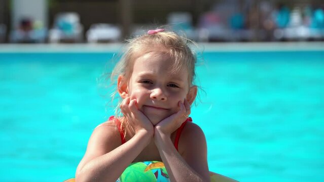 Portrait of little girl rests by hotel pool with blue water and inflatable swim ring and smiles. Summer vacation, holiday and travel