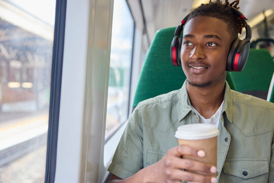 Young Man Commuting To Work Sitting On Train Wearing Wireless Headphones With Takeaway Coffee