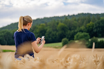Blonde junge Frau steht im erntereifen Weizenfeld und tippt auf ihrem Smartphone