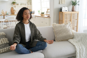 Serene mixed-race girl meditates at home, in lotus pose with mudra gesture on the sofa. Afro-haired, calm, practicing yoga. Healthy lifestyle, meditation.