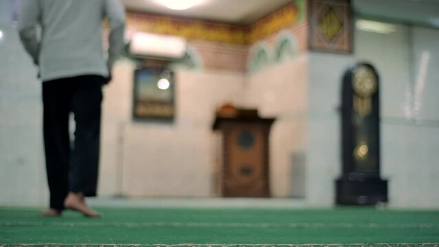 A man walks and then sits near the imam area of a mosque with a classic wall clock with a swinging pendulum and a pulpit in an antique Faletehan mosque in Jakarta. Taken with a medium shot and handhel