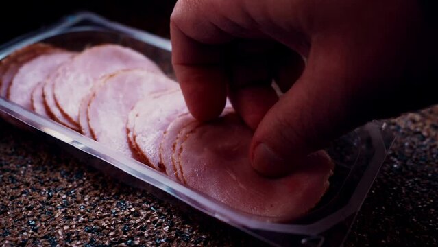 Caucasian man hand takes circle of thinly sliced ham from plastic package. Plastic packaging for chopped snacks on stone table. Sliced into thin pieces of ham on cutting serving board. Slow motion