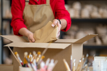 Woman worker of pottery shop packing order for client while standing by table against shelves.