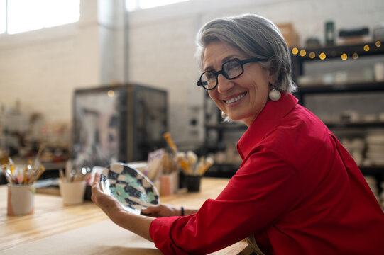 Woman potter in apron posing in ceramic workshop, holding crafted plate, looking at camera.