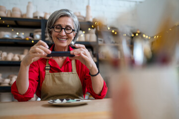 Pottery master woman photographing work result, using mobile phone for taking picture of earthenware