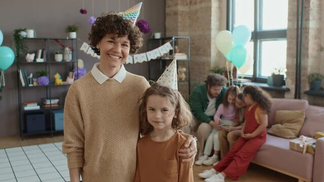 Medium portrait shot of young curly Caucasian woman in casual jumper and birthday party hat standing in living room hugging 9-year-old son and smiling while posing for photosMedium portrait shot of yo
