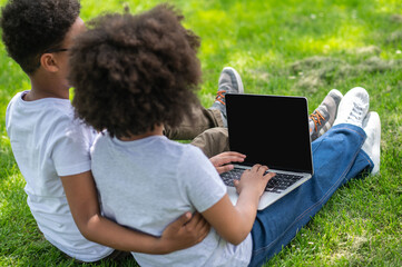 Children girl and boy with laptop sitting on grass watching movie browsing internet, blank screen.