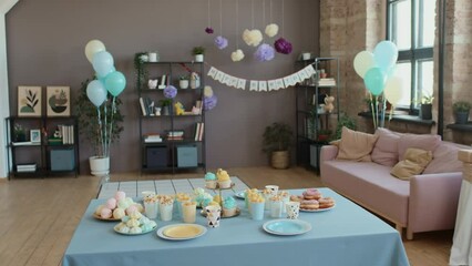 Full shot of table with sweet treats and snacks in middle of living room of family home, decorated with colourful balloons, pompoms and bunting, ready for kids birthday party before arrival of guests