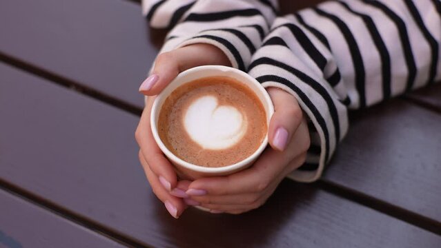 Closeup top view of unrecognizable young woman holding hot cup of coffee with heart shape. Close up cropped shot of unrecognizable female fingers holding cup of coffee with heart shaped latte art foam