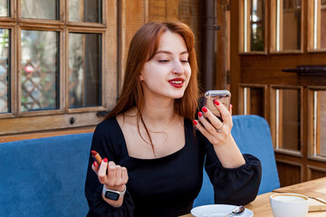 A girl in a cafe at a table paints her lips with red lipstick