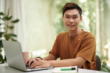 Cheerful Asian computer science student programming in laptop at home
