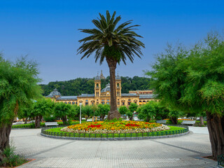 Obraz premium Alderdi Eder Gardens in San Sebastian, Spain, palm and tree–filled park by the beach, with colorful gardens. In the background the old city hall and colossal statue of Jesus overlooking the city.