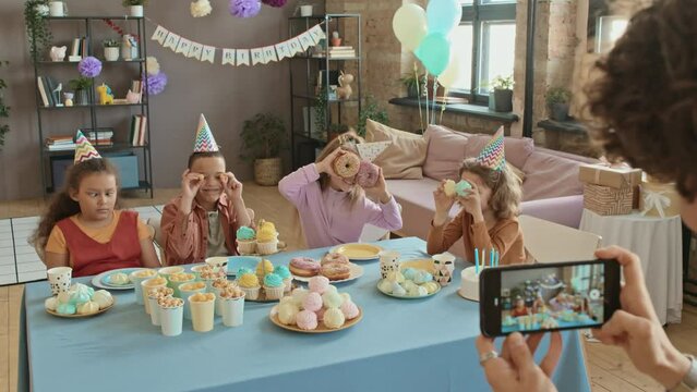 Over-shoulder shot of hands of unrecognizable woman holding smartphone and taking photos of diverse preteen children, sitting at table with sweets at birthday party, making silly faces, fooling around