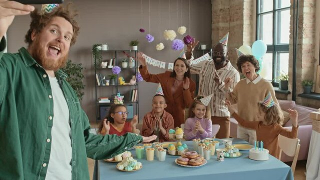 Medium shot of happy Caucasian man holding smartphone and taking group selfie of diverse parents and children at birthday party, all guests waving, cheering and clapping with excitement