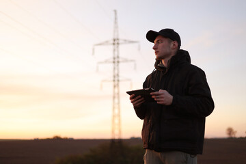 Young power engineer holds digital tablet against high voltage tower at field. Electrical engineer...