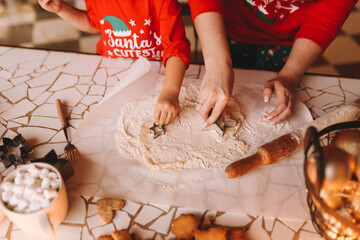 Mom and daughter in red pajamas are preparing for the holiday together preparing a festive meal in the kitchen. Human hands are cutting Christmas cookies out of dough. Selective focus
