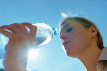 Young beautiful woman drinking pure water from a bottle on blue sunny sky background, focus on bottle