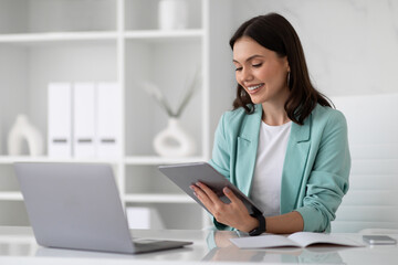 Smiling young european woman in suit at table with laptop typing on tablet in modern office interior