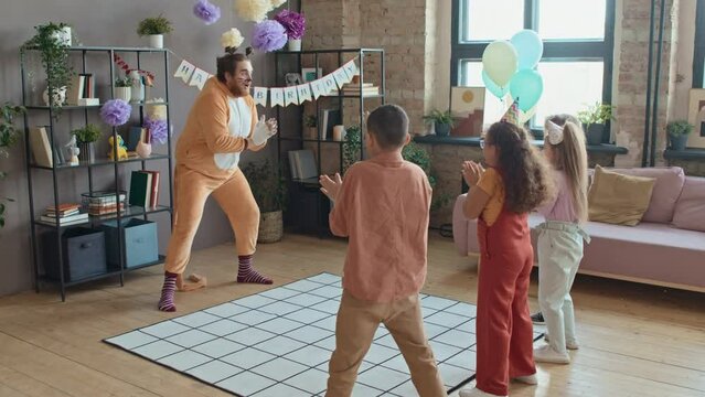 Full shot of male animator in cat character costume and make-up playing physical games with group of young children at home birthday party, clapping, marching and kids repeating
