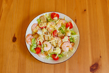 Fresh healthy caesar salad with prawns, cherry tomatoes, lettuce, parmesan cheese, dressing and croutons on wooden table. Top view