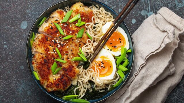 Asian noodles ramen soup with deep fried panko chicken fillet and boiled eggs in ceramic bowl with chop sticks and soy sauce on stone rustic table slowly rotating overhead . High quality 4k footage