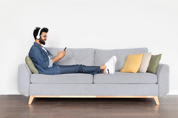 Relaxed young eastern guy chilling on couch with headphones, smartphone
