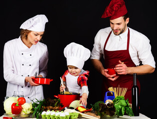 Happy family in chef uniform cooking at home. Family preparing healthy tasty food together in kitchen. Parents with child preparing vegetarian meal. Mother and father teaching little son how to cook.