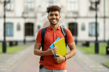 Cheerful Black Student Guy Holding Workbooks Posing With Backpack Near College Building