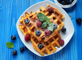 Waffles with blueberries and raspberries for breakfast on white plate over blue wooden table.