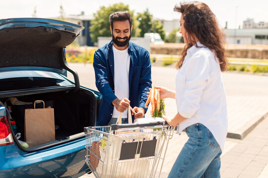 Young middle eastern spouses packing groceries into car trunk outdoors, standing on parking near shopping mall