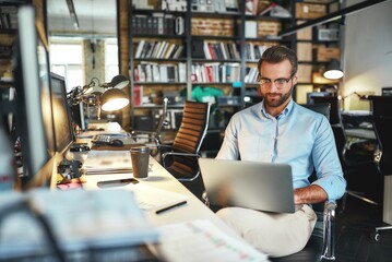 Comfortable workplace. Portrait of young and successful bearded man in eyeglasses working with laptop while sitting in modern office