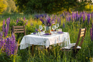 Romantic table decor for a loving couple on the blooming meadow with purple lupines. Two glasses of wine, flowers in a vase, silverware, fruits, wooden furniture and picnic basket. Sunset, golden hour