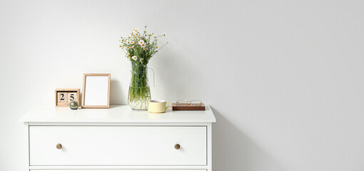 Vase with chamomiles and decor on chest drawers near light wall. Banner for design