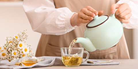 Woman pouring natural chamomile tea from teapot into cup on table in kitchen