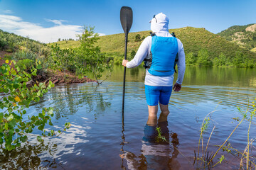 male stand up paddler wearing sun hat and life jacket is standing in water on lake shore -...