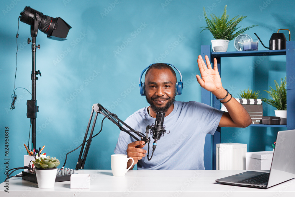 Poster smiling blogger in headphones waving hi and speaking in microphone while live streaming for social m