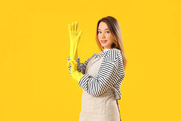 Young woman putting rubber gloves on yellow background