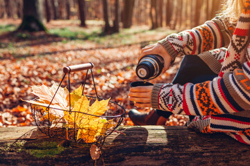 Woman pouring tea from thermos into cup relaxing in autumn forest sitting on trunk. Close up of drink and yellow leaves - obrazy, fototapety, plakaty