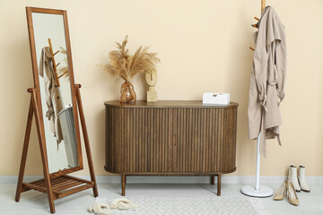 Chest of drawers, mirror and rack with coat near beige wall in hall