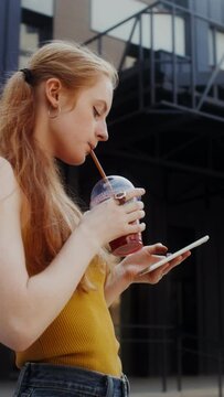 A young red-haired woman uses a mobile phone and drinks juice from a disposable cup with a straw while walking on a city street