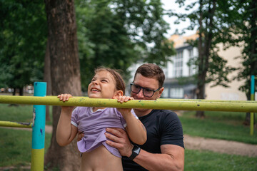 Obraz premium Father with child girl doing pull-ups on workout outdoor area. Healthy active lifestyle, happy family time. Modern fatherhood concept 