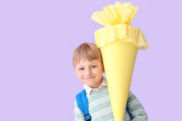 Happy little boy with backpack and yellow school cone on lilac background