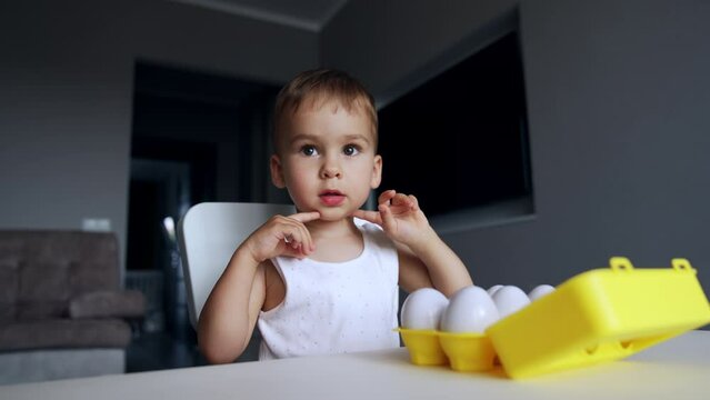Baby boy sitting at desk plays with toy eggs. Kid opens and closes the box with eggs with interest.