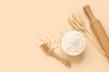Bowl with wheat flour, spikelets, rolling pin and scoop on beige background