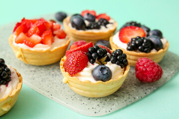 Board of tasty tartlets with whipped cream and berries on turquoise background