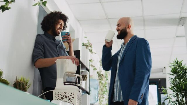 Colleagues laughing on coffee break, enjoying conversation and joking around in open space shared office. Coworkers relaxing at work, talking to each other in modern workplace.