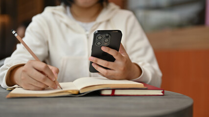 A young Asian female college student using her smartphone while doing her homework