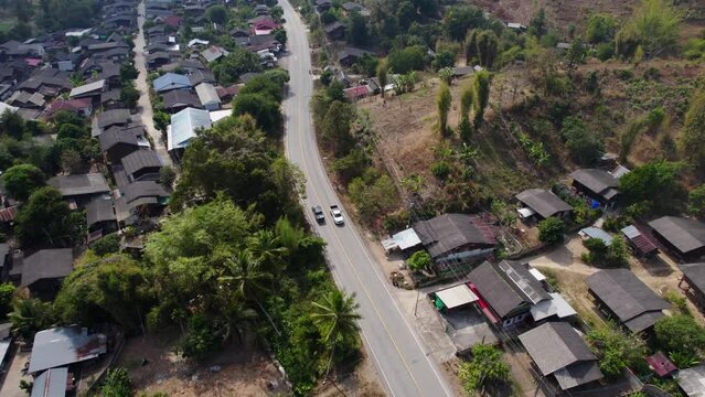 Village in Thailand filmed from above. The camera following the car passing by the road. Misty mountains in the background. Palm trees on the foreground.