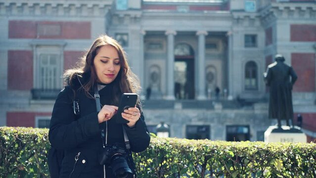 Pensive young Latin woman in warm clothes with professional photo camera using smartphone while standing near Prado Museum in Madrid, Spain