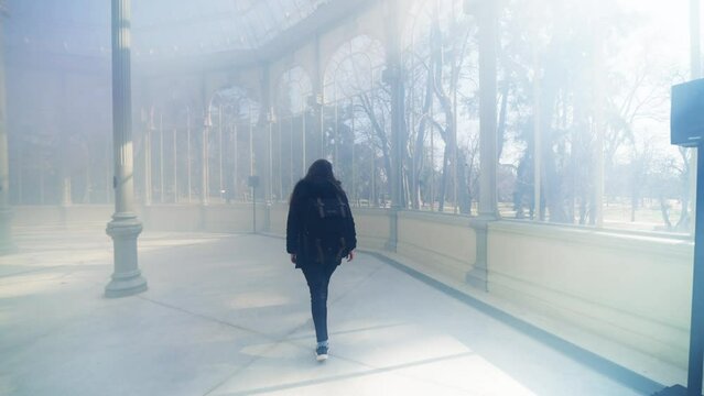 Young Latin woman tourist in warm clothes with professional photo camera walking through the mist and the rays of light and standing inside of The Glass Palace in Madrid, Spain 